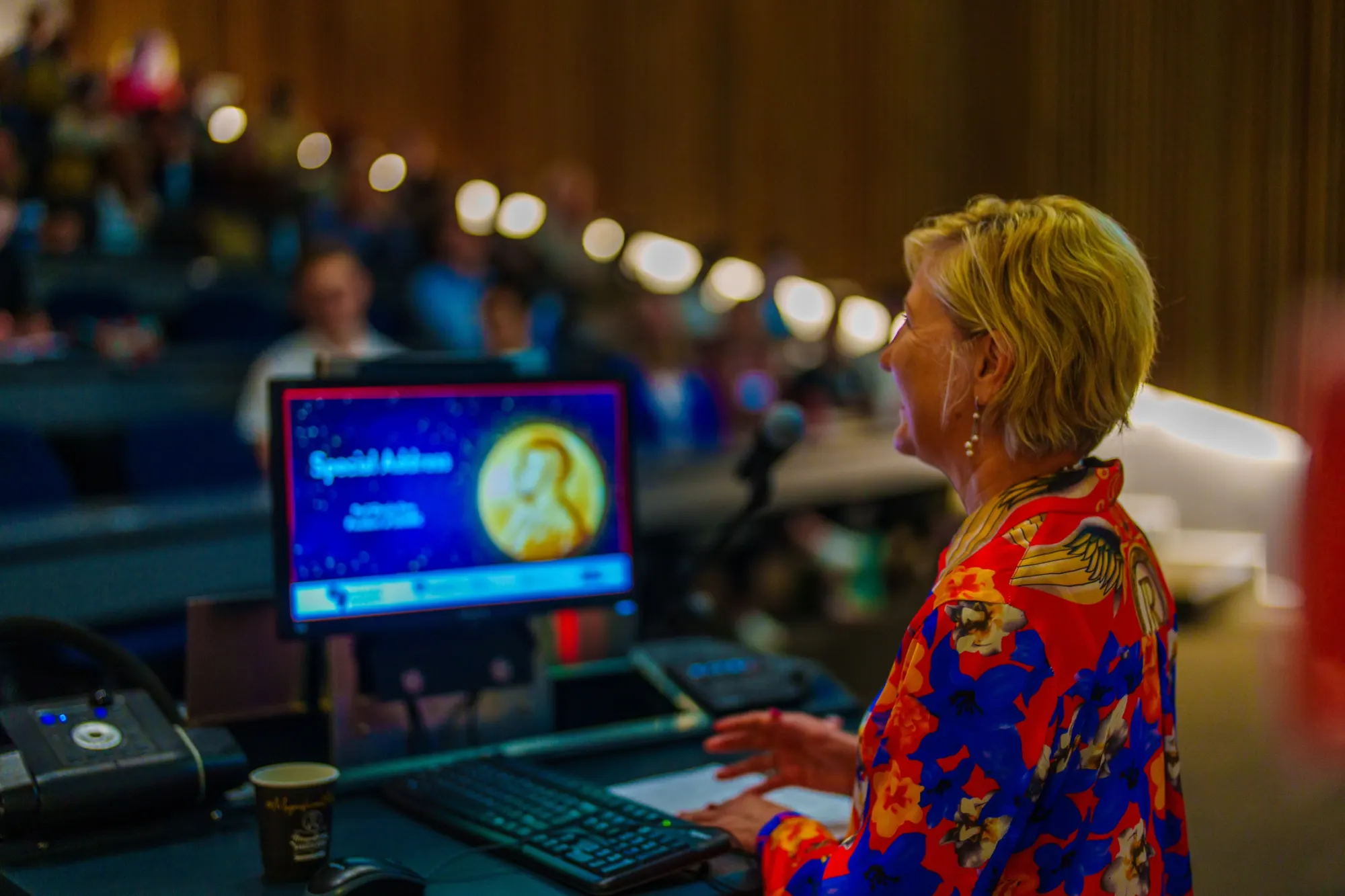 A host at the lectern of a lecture theatre as the Nobel Laureate special address slide shows on screen