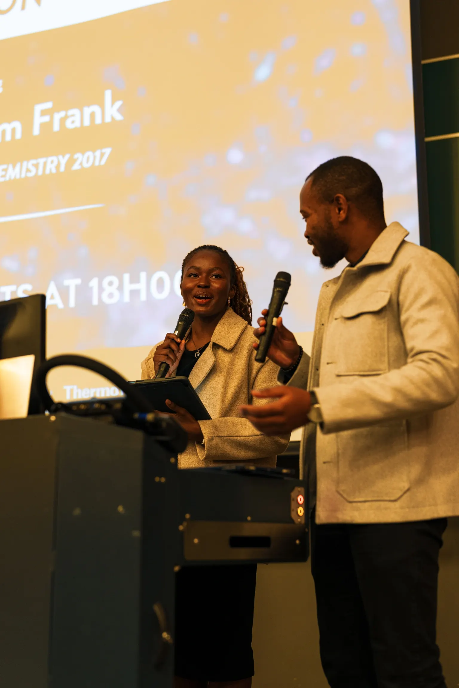 Two ARS hosts with microphones on stage in front of the Nobel Laureate event slide